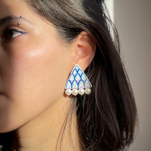 May include: A triangular earring featuring a white base with a blue and pink scalloped pattern. The bottom edge of the triangle is adorned with four small, round, white pearls arranged in a horizontal row along the base of the earring.