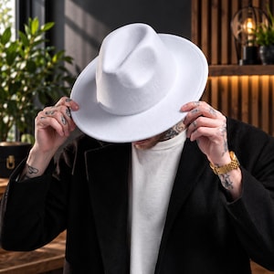 May include: A solid white fedora hat with a structured crown and a wide, flat brim. The hat features a smooth, uniform texture and a matching white band around the base of the crown, providing a clean and classic look for any headwear collection.
