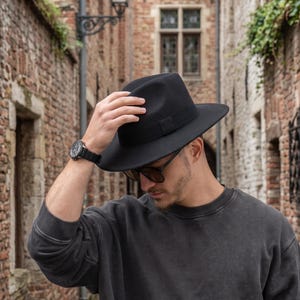 May include: A black fedora hat with a wide brim and a matching black ribbon band, paired with a dark grey long-sleeved crewneck sweatshirt, a black wristwatch with a link band, and dark-tinted sunglasses with a thin frame.