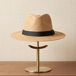 Op de afbeelding: Een beige strooien fedora hoed met een brede rand en een zwarte grosgrain lintband. De hoed heeft een ingedeukte kroon en een geweven textuur, weergegeven op een goudkleurige metalen standaard met een ronde voet en een gebogen steun.