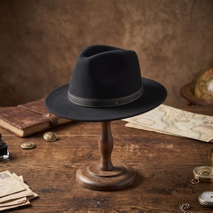 May include: A black fedora hat with a structured crown and a wide brim, featuring a dark brown leather band around the base. The band is detailed with visible stitching and small metal eyelets, providing a contrast to the solid black felt material.