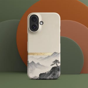 May include: A beige smartphone case featuring a black and white ink wash landscape illustration of misty mountains and a single tree. A horizontal gold foil band separates the upper solid beige section from the detailed mountain scene below.