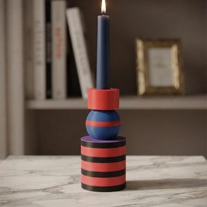 May include: A lit dark blue taper candle sits in a stacked candle holder. The holder features a red ring at the top, a blue and red sphere in the middle, and a cylindrical base with alternating horizontal red and black stripes throughout the piece.