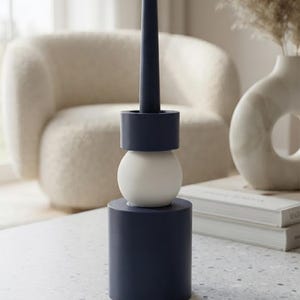 May include: A dark blue taper candle stands inside a stacked candle holder. The holder consists of a large dark blue cylinder base, a smooth white sphere in the center, and a smaller dark blue cylinder at the top to secure the candle in place.