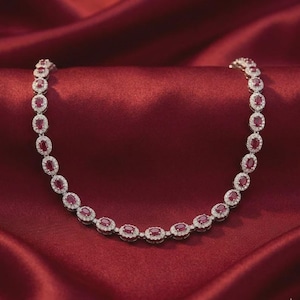 May include: A necklace featuring a series of oval-shaped red gemstones, each surrounded by a halo of small, clear, sparkling stones. The links are connected in a continuous chain, creating a uniform design that displays a vibrant red and silver colour palette.