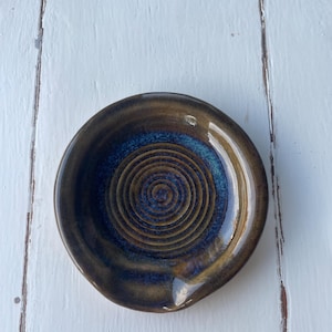 May include: A round ceramic spoon rest featuring a glossy glaze with a deep blue centre and a brown outer rim. The interior surface displays a raised concentric spiral pattern, and the edge includes a small indentation for resting a utensil.