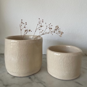 May include: Two cylindrical ceramic vessels with a glossy, off-white crackle glaze finish. The taller vessel on the left contains a small sprig of dried baby's breath flowers. Both containers feature a smooth, rounded rim and a solid base.