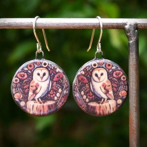May include: A pair of round dangle earrings featuring a detailed illustration of a barn owl perched on a tree stump, surrounded by a floral pattern in shades of brown, red, and cream, suspended from silver-toned metal ear wires.