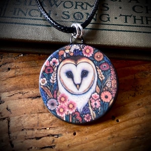 May include: A circular pendant necklace featuring a detailed illustration of a white barn owl surrounded by a colorful floral wreath of pink, orange, and blue flowers. The pendant hangs from a black cord and displays a vibrant, artistic design.