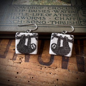 May include: A pair of square white earrings featuring a black ink illustration of a sloth hanging from a tree branch. Each earring is attached to a silver-toned metal hook for hanging. The design is identical on both pieces, showing the sloth in profile.