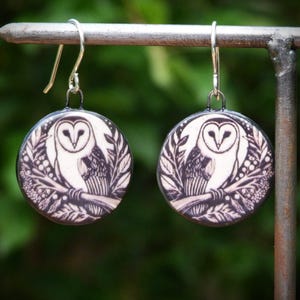 May include: A pair of circular drop earrings featuring a black and white illustration of a barn owl perched on a branch surrounded by foliage. The earrings have silver-toned metal hooks and a smooth, round surface with a detailed graphic design.