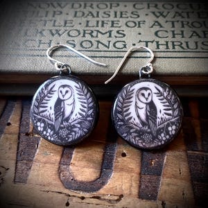 May include: A pair of round drop earrings featuring a black and white illustration of a barn owl perched on a branch surrounded by leaves and berries. The earrings have silver-toned metal hooks and a glossy finish on the circular pendant faces.