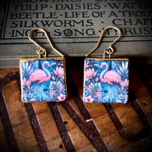 May include: A pair of square drop earrings featuring a vibrant print of pink flamingos surrounded by blue and pink tropical foliage. The earrings have gold-toned metal edges and are suspended from thin, curved gold-toned metal ear wires.