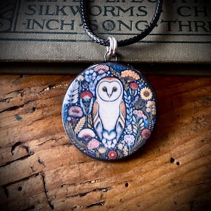 May include: A circular pendant necklace featuring a detailed illustration of a white barn owl surrounded by colorful wildflowers and foliage. The pendant hangs from a black braided cord, showcasing a vibrant botanical design with a dark blue background.
