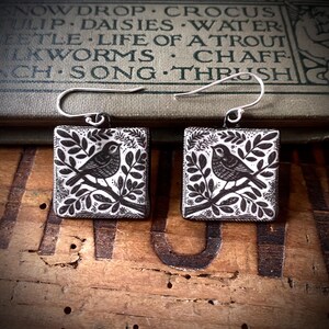 May include: A pair of square drop earrings featuring a black and white illustration of a bird perched on a leafy branch. The earrings have silver-toned metal hooks and a flat, square-shaped pendant design with a detailed botanical pattern.