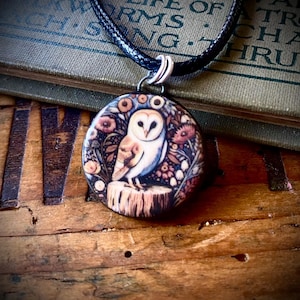 May include: A round pendant necklace featuring a barn owl perched on a tree stump, surrounded by a floral pattern in shades of brown, cream, and muted red. The pendant hangs from a black cord and is secured by a silver-toned metal bail.
