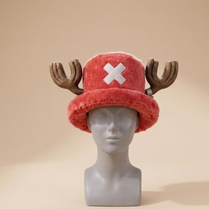 Puede incluir: Un sombrero de pescador de felpa color rosa coral con un emblema de cruz blanca en la parte delantera y dos grandes astas marrones texturizadas que sobresalen de los lados. El sombrero se exhibe sobre una cabeza de maniquí de tono neutro.
