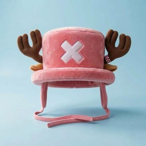 Op de afbeelding: Een pluche roze hoed met twee grote bruine geweien die aan de zijkanten uitsteken en een wit kruissymbool in het midden aan de voorzijde. De hoed heeft een brede rand en twee lange stoffen banden die aan de zijkanten naar beneden hangen.