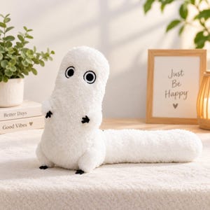 Puede incluir: Un juguete de peluche blanco con un cuerpo largo y cilíndrico y una cabeza redondeada. Presenta dos ojos grandes, circulares, negros y blancos, y pequeñas manos y pies negros de tres dedos. La textura del peluche es suave y esponjosa.