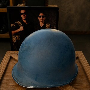 Könnte beinhalten: Ein matter, blauer Helm im Militärstil mit einer glatten, abgerundeten Kuppelform und einer breiten, geschwungenen Krempe. Die Oberfläche des Helms weist ein durchgehendes, einfarbig blaues Finish mit subtilen Texturvariationen auf.