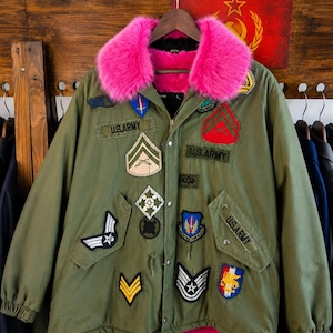 May include: An olive green military-style field jacket featuring a vibrant hot pink faux fur collar and lining. The exterior is decorated with numerous embroidered patches, including several U.S. Army insignia, chevron stripes, and various colorful emblems.