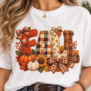 May include: A white t-shirt featuring a graphic of the word FALL in large, textured letters. The letters are decorated with plaid, floral, and solid patterns, accented by pumpkins, a sunflower, autumn leaves, berries, bows, and buttons.