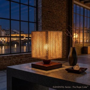May include: A square table lamp featuring a shade wrapped in natural tan rope, set on a dark wooden base. The lamp emits a warm glow. Next to it sits a dark vase with thin branches and a small silver pocket watch in a dark tray. Text reads ELEMENTAL Series - The Rope Cube.
