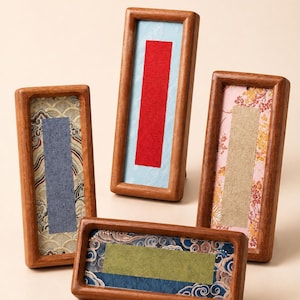 May include: Four rectangular wooden frames with rounded edges, each containing a unique fabric insert. The inserts feature patterned backgrounds in gold, light blue, pink, and dark blue, with solid-colored rectangular patches in gray, red, beige, and olive green.
