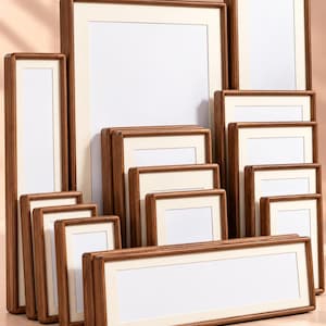 Puede incluir: Una colección de marcos de fotos rectangulares de madera en varios tamaños, cada uno con un acabado de grano de madera marrón natural y un borde grueso de cartón color crema que rodea un espacio central blanco vacío para fotos o arte.