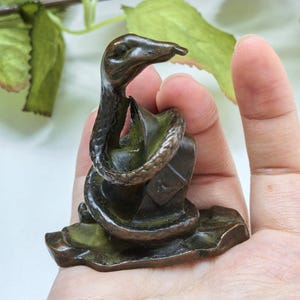 Könnte beinhalten: Eine kleine, dunkel bronzefarbene Metallfigur, die eine Schlange zeigt, welche sich um einen rechteckigen Block windet. Die Schlange hat eine texturierte, geschuppte Oberfläche und ist mit erhobenem Kopf dargestellt. Sie ruht auf einer flachen Basis.