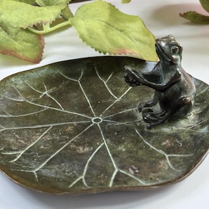 Könnte beinhalten: Eine dunkel bronzefarbene Metallschale in Form eines Seerosenblatts mit einer kleinen, skulptierten Froschfigur am Rand. Die Schale weist eingravierte weiße Aderdetails auf der Oberfläche auf und der Frosch ist in sitzender Position dargestellt.