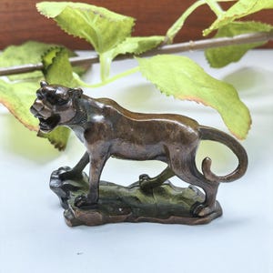 Könnte beinhalten: Eine kleine, dunkel bronzefarbene Metallfigur eines schleichenden Panthers mit geöffnetem Maul, die auf einem strukturierten, felsigen Sockel steht. Der Panther ist in einer geduckten Haltung mit nach oben gebogenem Schwanz dargestellt.