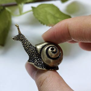 Puede incluir: Una pequeña figura de caracol hecha de metal en tono latón. El caracol presenta una superficie muy texturizada y rugosa en el cuello y la cabeza, que contrasta con una concha lisa en espiral con un punto central oscuro y hundido.