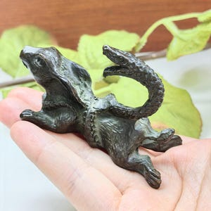 May include: A dark bronze-toned metal figurine depicting a rabbit with a snake coiled around its midsection. The rabbit is shown in a dynamic, leaping pose with its ears laid back, while the snake's head is raised and curved upwards.
