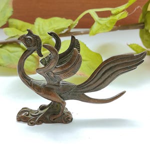 Pode incluir: Uma pequena estatueta de metal na cor bronze escuro representando uma ave mítica com um pescoço alongado, uma crista na cabeça e grandes asas abertas. A criatura está em posição ereta sobre uma base com detalhes esculpidos em espiral.