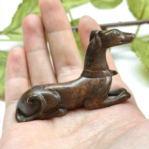 Könnte beinhalten: Eine kleine, dunkelbraune Metallfigur eines Hundes in ruhender, liegender Position. Der Hund hat eine glatte, verwitterte Textur, ein Halsband und einen eingerollten Schwanz nahe der Hinterhand. Die Figur ist aus massivem Metall gegossen.