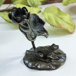 Könnte beinhalten: Eine dunkel bronzefarbene Metallskulptur, die einen Frosch auf einem großen Seerosenblatt zeigt. Eine einzelne Lotusblüte sitzt auf einem hohen, geschwungenen Stiel, der aus der Basis ragt, neben einem großen, aufrechten Seerosenblatt hinter dem Frosch.