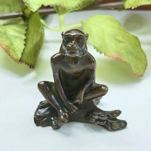 May include: A small, dark brown bronze figurine of a monkey sitting in a relaxed, forward-facing posture on a textured, log-like base. The monkey has detailed facial features, defined limbs, and a smooth, metallic finish throughout the sculpture.