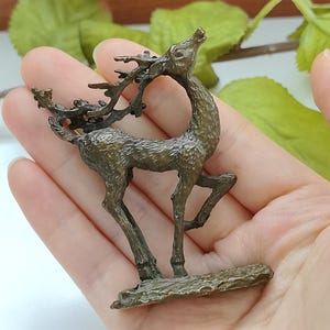 May include: A bronze-toned metal deer figurine standing on a rectangular base. The deer is depicted in a dynamic pose with its head tilted upward and large, textured antlers extending backward. The surface has a weathered, dark metallic finish.