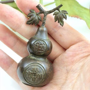 May include: A dark bronze-toned metal gourd-shaped vessel featuring a stem with two leaves at the top. The body displays two circular embossed medallions, with the lower medallion featuring Chinese characters. The surface has a weathered patina.