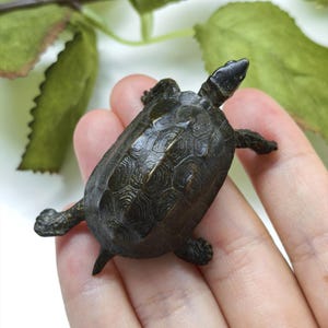 Puede incluir: Una pequeña figura de tortuga de metal en tono bronce oscuro que presenta un caparazón texturizado detallado con un patrón geométrico. La tortuga se muestra en posición de gateo con la cabeza extendida hacia adelante y las cuatro patas extendidas.