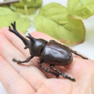 以下が含まれることがあります： 頭部に目立つ二股の角、質感のあるセグメント化された外骨格、6本の細い脚を備えたダークブラウンのメタリックなカブトムシの置物で、詳細でリアルな彫刻のフォルムで表現されています。