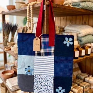 May include: A patchwork tote bag featuring various blue and white fabric squares with polka dot, striped, and floral patterns. The bag has two red fabric handles and a brown paper tag attached with string that displays the text £20 in black ink.