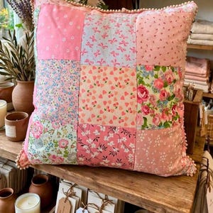 May include: A square throw cushion featuring a patchwork design of nine distinct fabric squares in shades of pink, white, and blue. The patterns include floral prints, small cherries, and bows. The edges are finished with a small pom-pom trim.