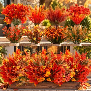 May include: A collection of artificial autumn-themed floral arrangements featuring vibrant red, orange, and yellow maple leaves, ferns, and small daisy-like flowers. The display includes individual potted plants and a large rectangular wooden planter.