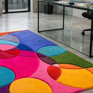 May include: A rectangular area rug featuring a vibrant, multi-colored geometric pattern of overlapping circles and shapes in shades of bright pink, orange, yellow, red, blue, purple, and green, with a soft, plush texture covering the floor surface.