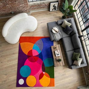 May include: A rectangular area rug featuring a vibrant geometric pattern of overlapping circles in shades of bright pink, orange, purple, blue, and green. The rug is positioned in a living room with a white curved sofa and a gray sectional sofa.