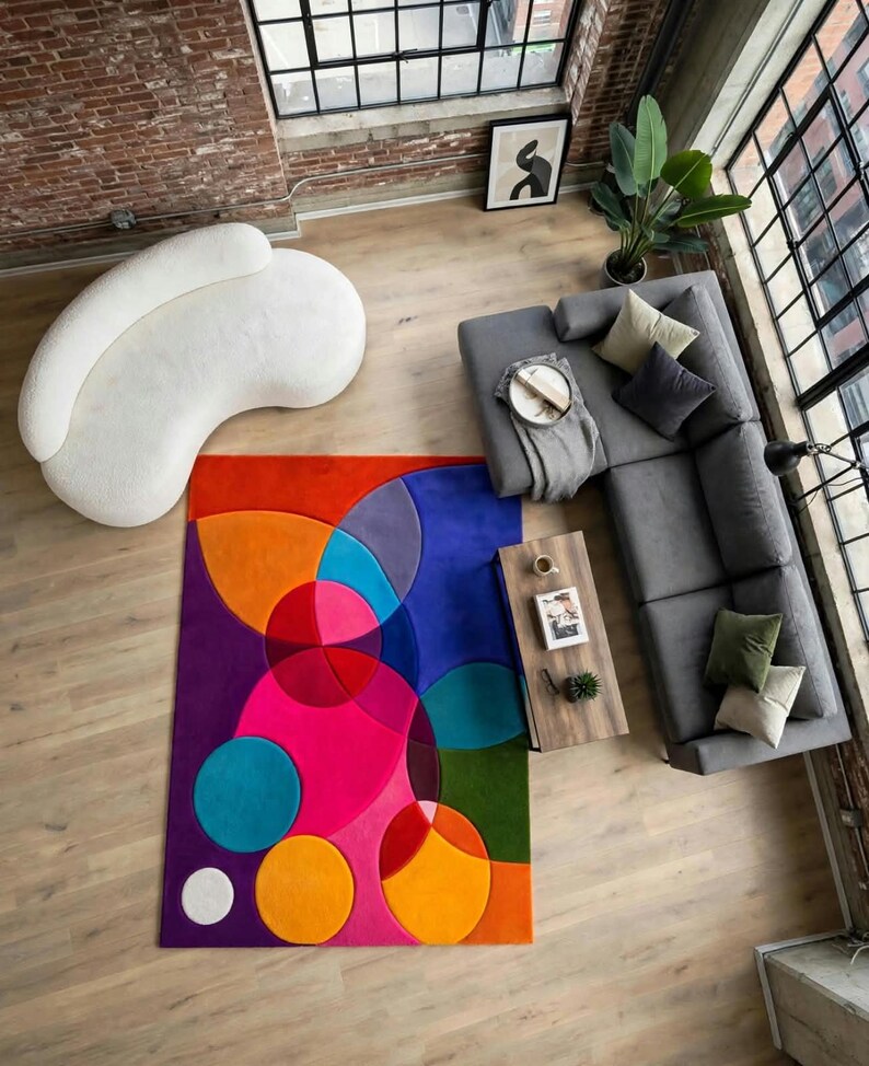May include: A rectangular area rug featuring a vibrant geometric pattern of overlapping circles in shades of bright pink, orange, purple, blue, and green. The rug is positioned in a living room with a white curved sofa and a gray sectional sofa.