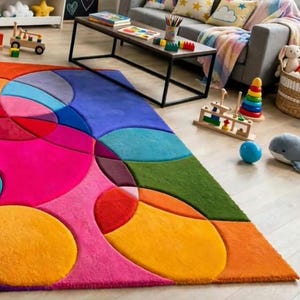 May include: A rectangular area rug featuring a vibrant geometric pattern of large, overlapping circles and curved shapes in bright shades of pink, orange, yellow, green, blue, and purple, with a soft, plush texture covering the floor surface.