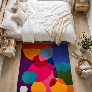May include: A rectangular area rug featuring a vibrant geometric pattern of overlapping circles and semi-circles in shades of magenta, purple, orange, blue, red, and green, placed on a light-colored wooden floor in a bedroom setting.